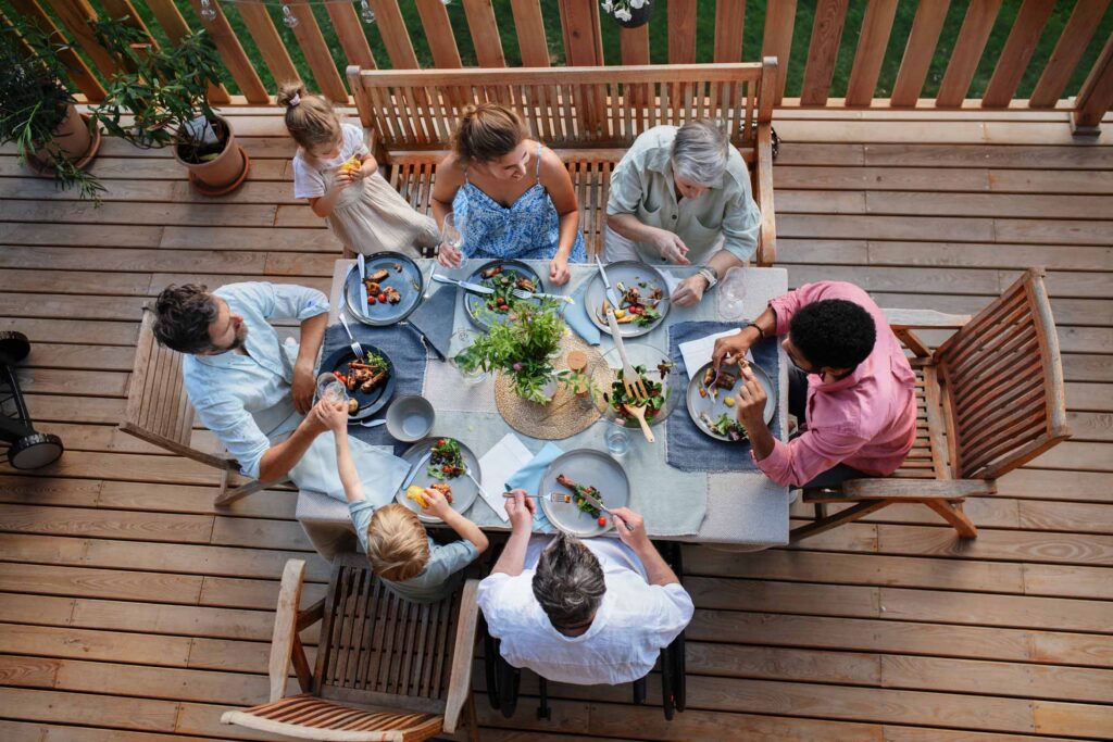 Top View Of 3 Generations Family Eating At Barbecue Party Dinner On Patio, People Sitting At Table On Patio With Grill.